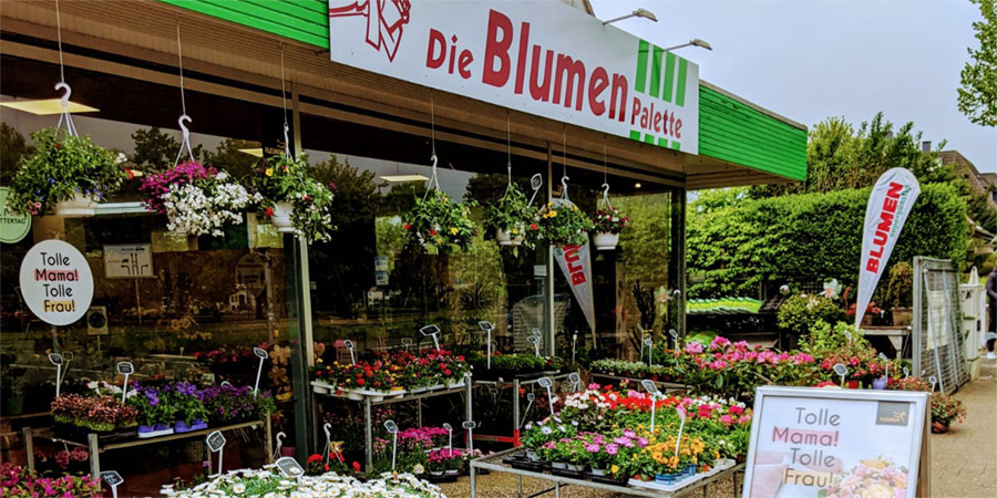 Schau mal, ein farbenfrohes Blumenfachgeschäft! "Die Blumen Palette" lockt mit hängenden Blumenampeln und liebevollen Muttertags-Angeboten. Einfach toll!